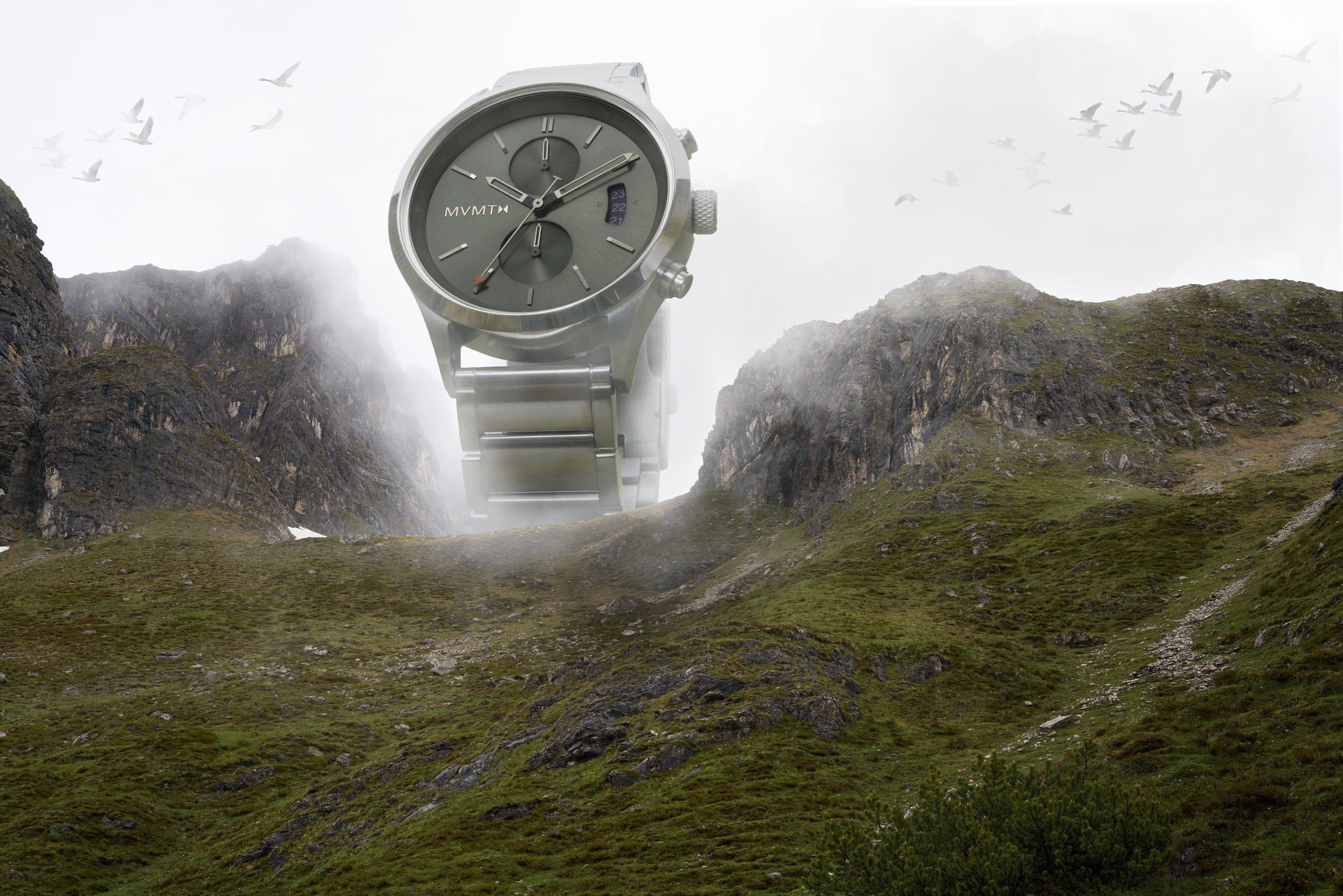 An oversized silver chronograph rising from a misty mountain valley with birds overhead