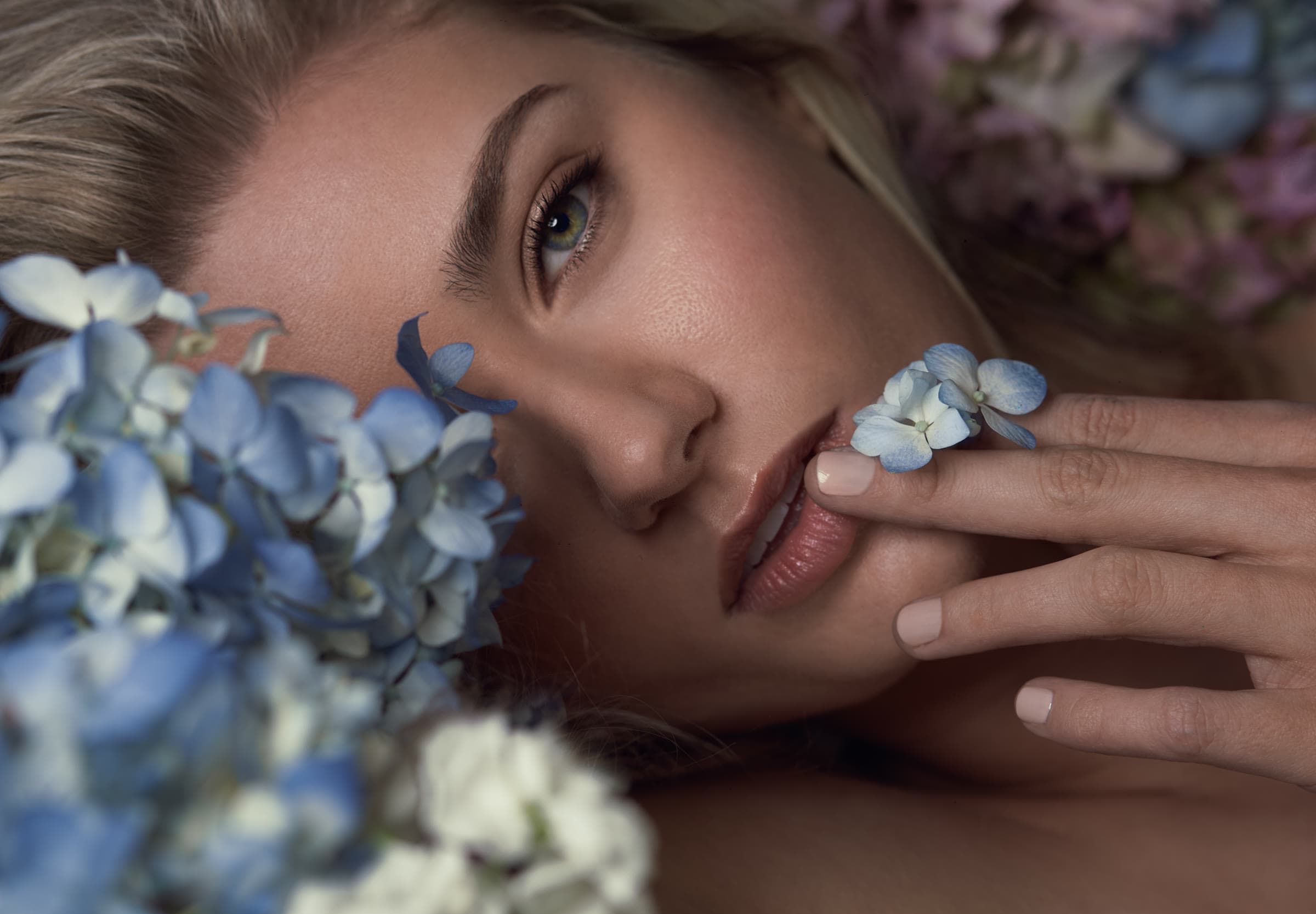 A blonde model among blue hydrangeas, a blossom held at her lip